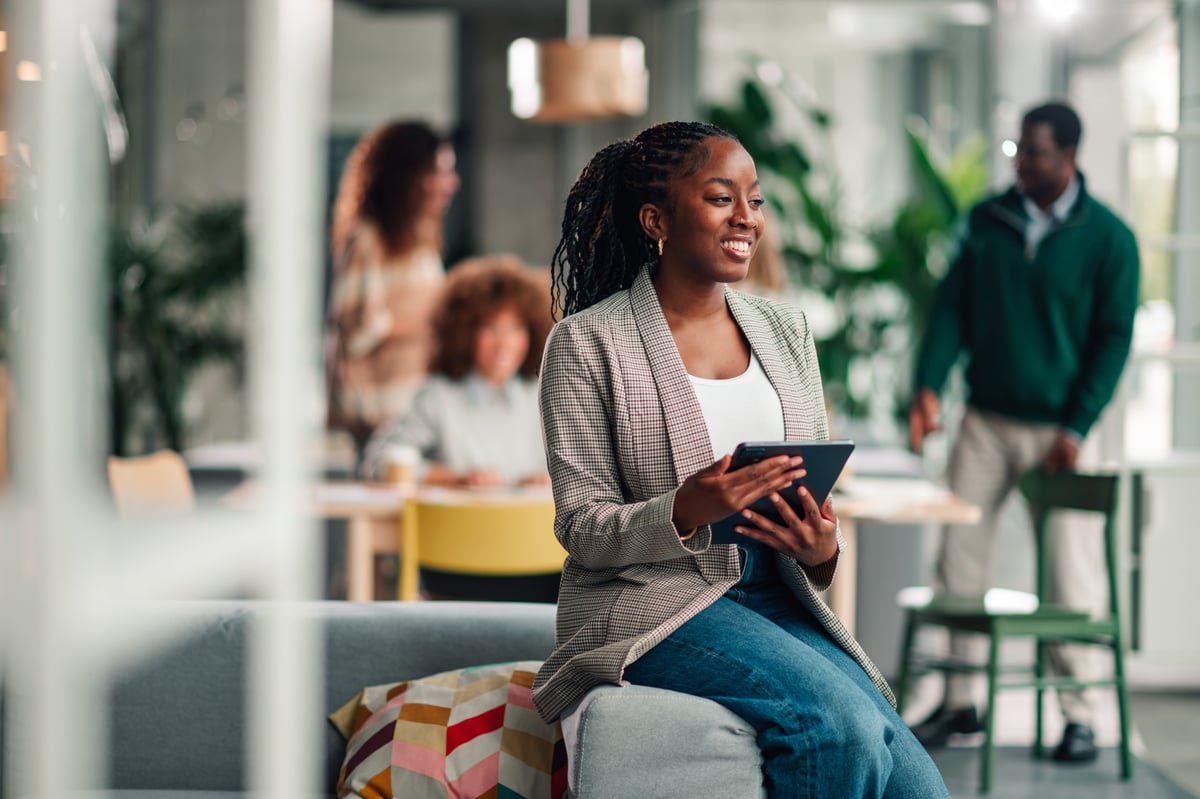Young adult businesswoman smiling, using a digital tablet while sitting in a contemporary office space