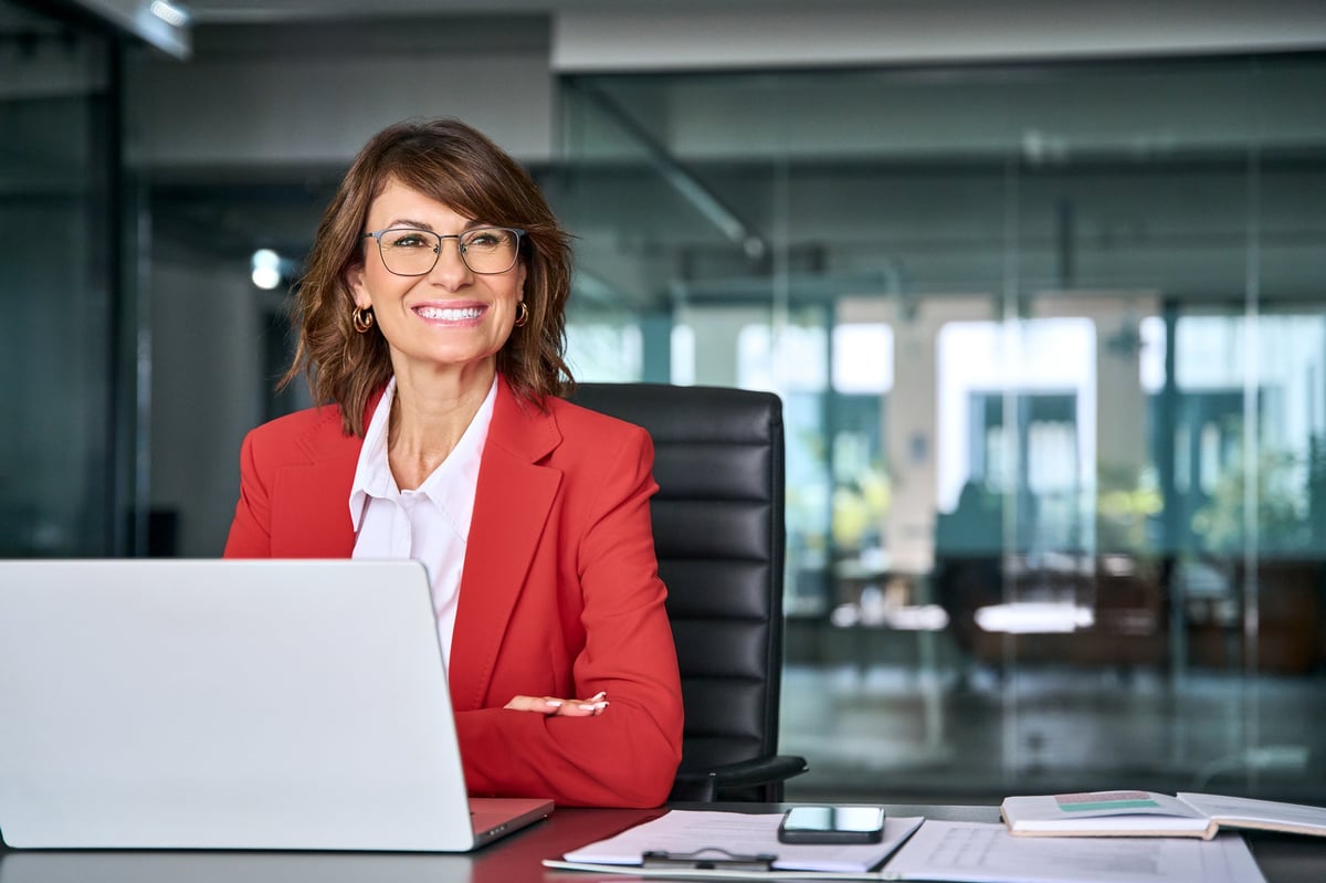 Smiling middle aged business woman using laptop at work office, professional female executive entrepreneur in red suit