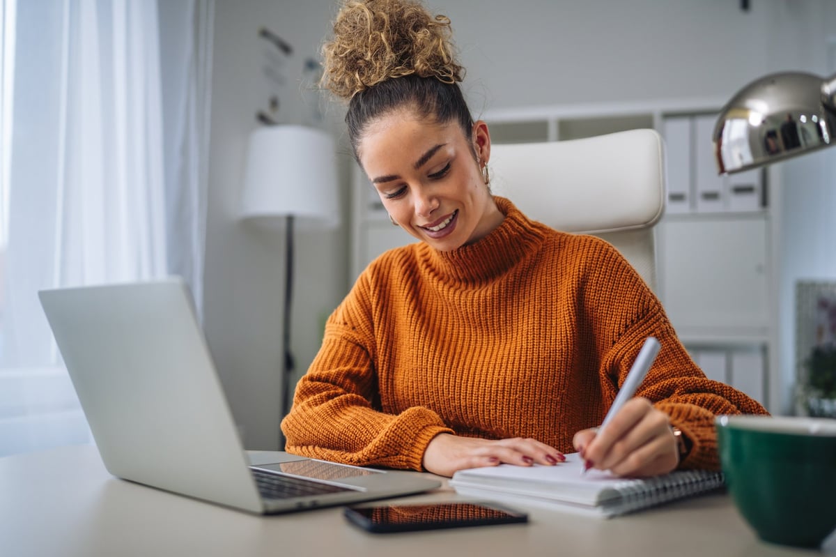 Young professional woman with curly hair bun wearing an orange sweater sitting at her desk, typing on a laptop, and taking notes in a spiral notebook