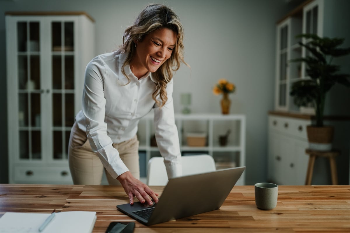 Young professional woman leaning over a laptop at a wooden desk, smiling while working from a bright home office