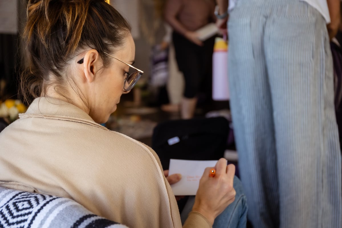 Woman wearing glasses writing or taking notes on a clipboard at an indoor event with people in the background