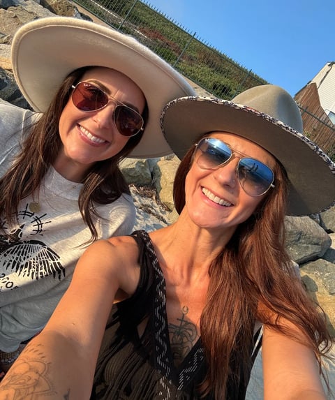Two women wearing cowboy hats and sunglasses smiling at the camera outdoors on a sunny day