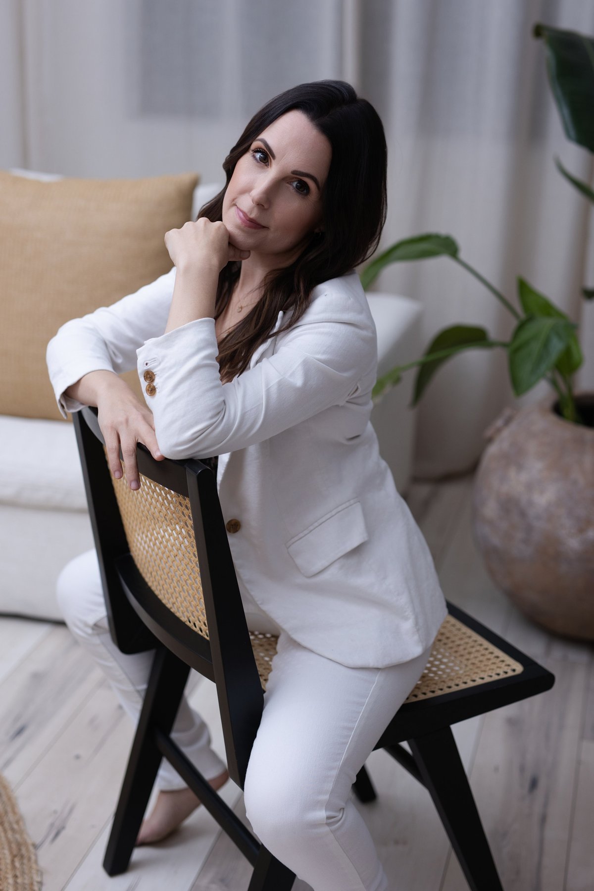 Woman in white blazer and pants sitting on black chair with hand to face, potted plants in background