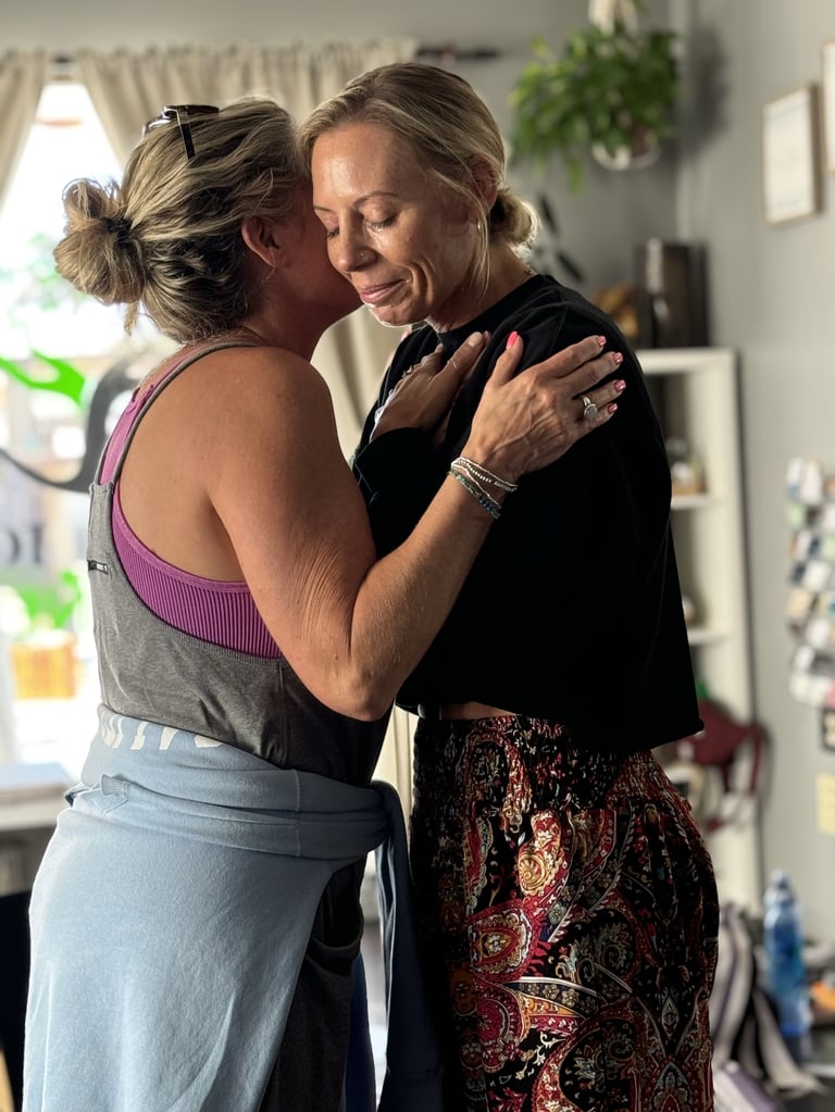 Two women embracing and smiling in a bright kitchen, one wearing a purple sports bra and the other in black top with patterned skirt