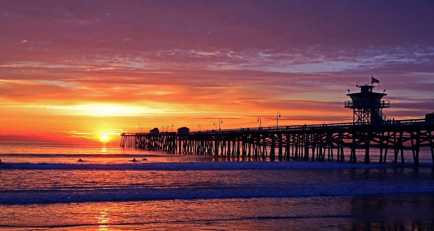 Vibrant sunset over the ocean with a pier extending into the water, glowing orange and purple sky reflecting on the sea