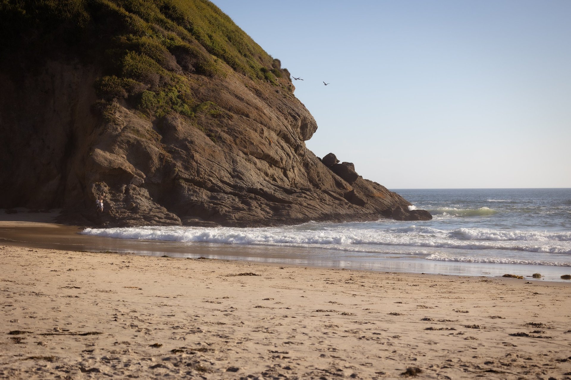 Rocky coastal cliff overlooking a sandy beach with waves breaking on the shore under a clear blue sky