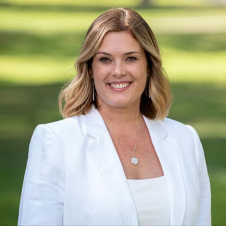 Professional headshot of a smiling woman with shoulder-length blonde hair wearing a white blazer and pendant necklace against a blurred green background