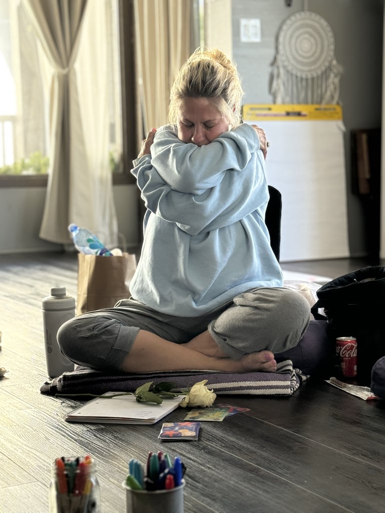 Woman sitting on floor hugging herself with art supplies scattered around in bright bedroom studio space