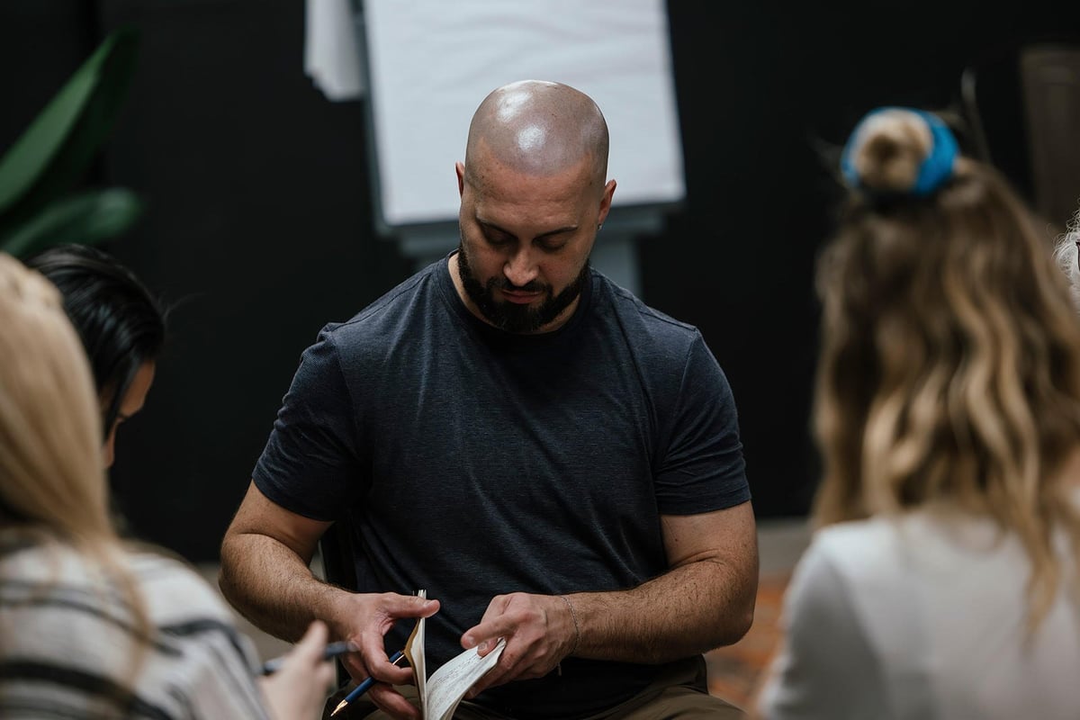 Bald man in dark t-shirt speaking to a small group of people in an indoor workshop or classroom setting