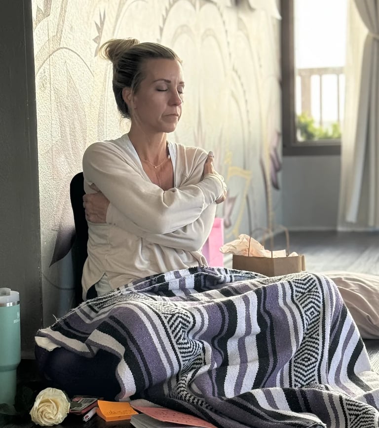 Woman sitting on bed against wall with arms crossed, wearing cream cardigan over patterned blanket, looking thoughtful by window
