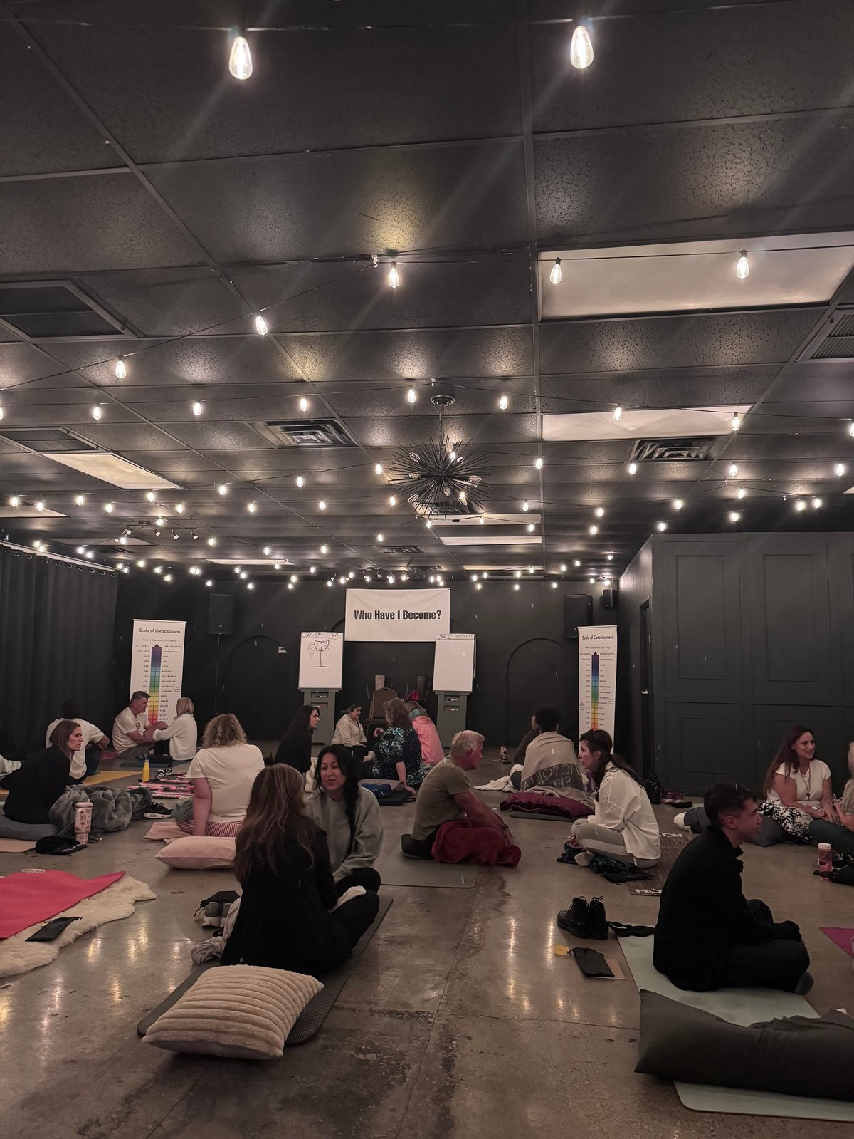 Group of people sitting on yoga mats in a dimly lit studio with string lights and a sign reading Who Move I Become