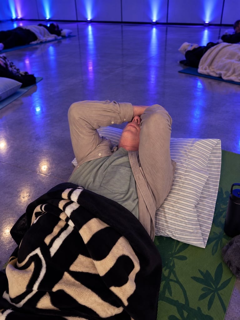 Person lying on yoga mat during relaxation session in dimly lit room with blue lighting and other participants resting nearby