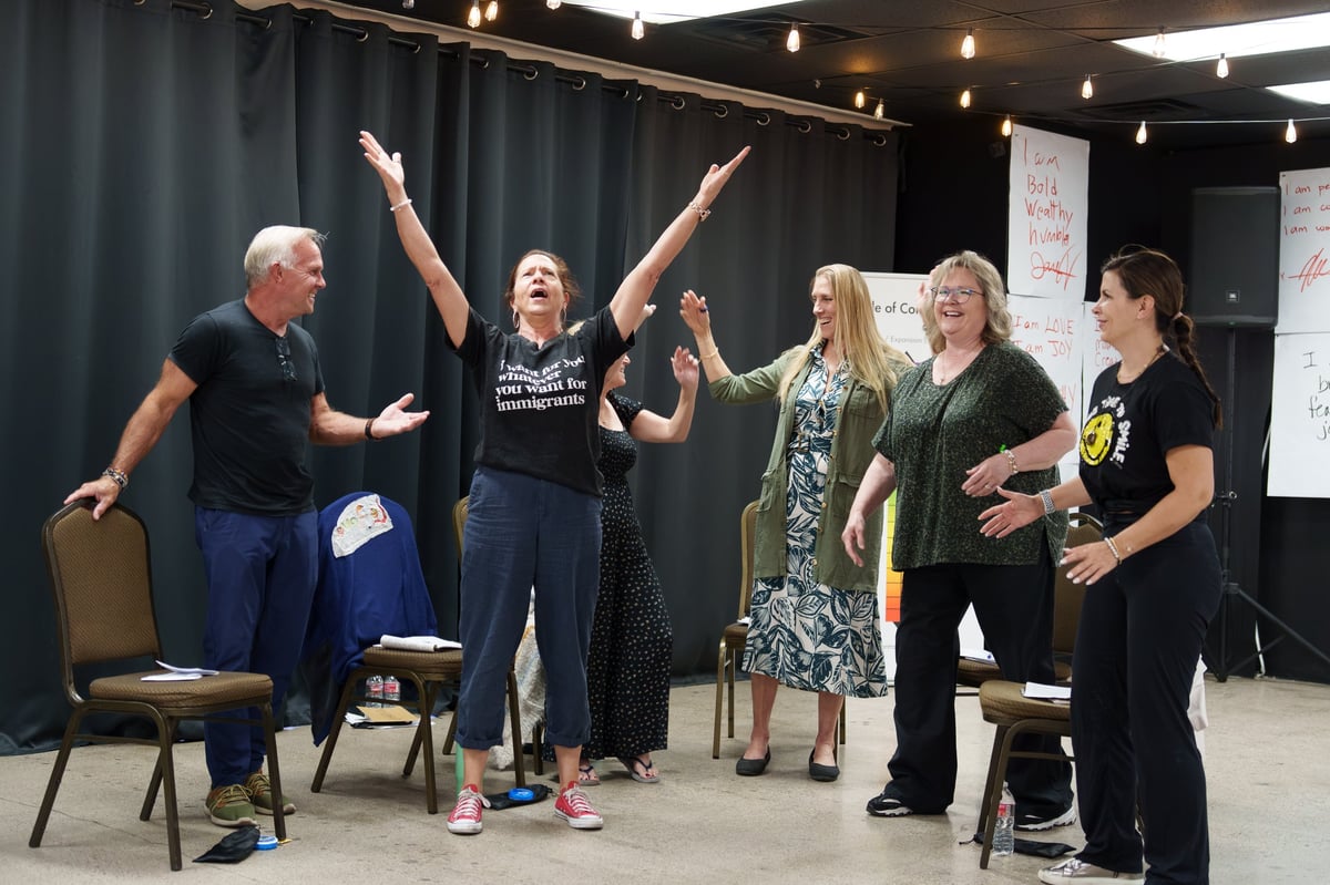 Group of six people in a workshop setting, with a woman in the center raising both arms in celebration while others stand nearby smiling