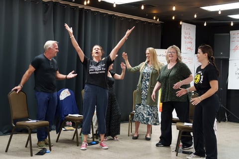 Group of six people in a workshop setting, with a woman in the center raising both arms in celebration while others stand nearby smiling