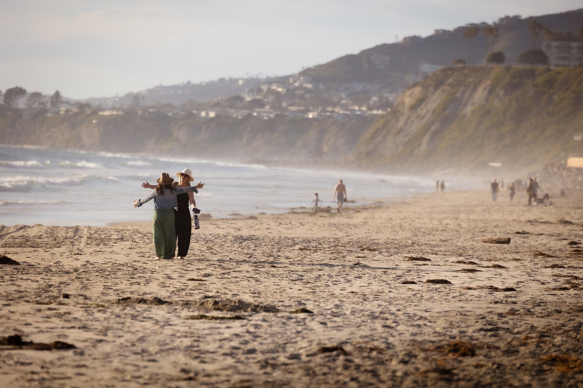 Surfers walking out of ocean onto sandy beach with cliffs and houses visible in background