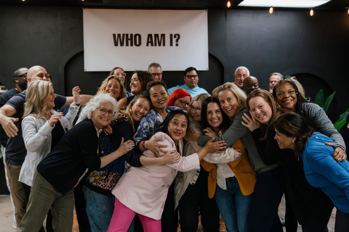 Group of diverse people gathered together under a WHO AM I? sign, smiling and posing for a team photo indoors.