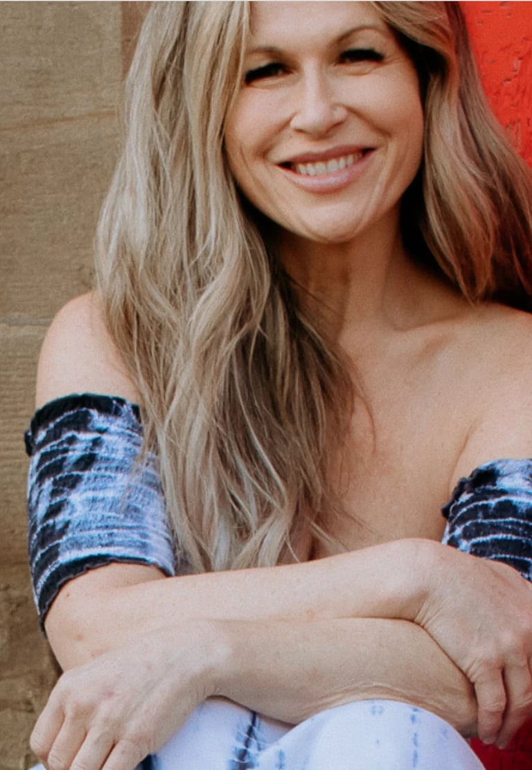 Woman with long blonde hair wearing blue tie-dye sleeves, smiling at camera against red and tan background