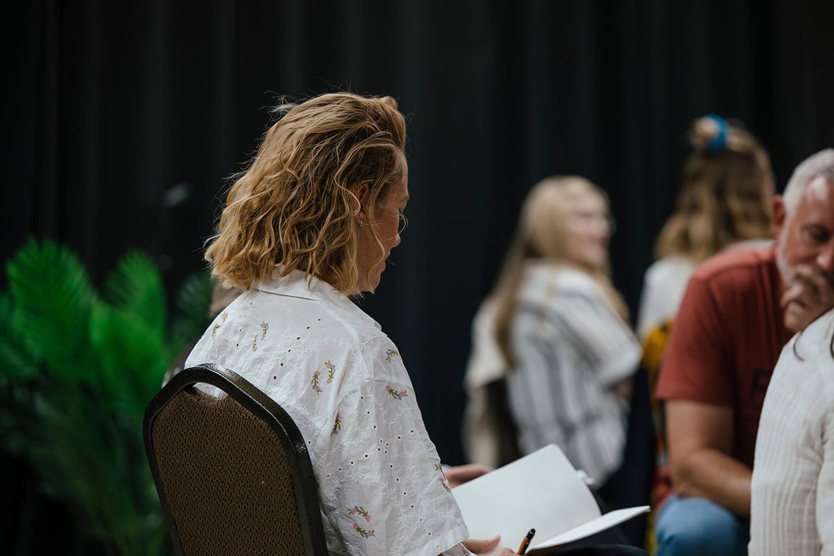 Woman with wavy blonde hair sitting in chair holding a paper, surrounded by other people at an indoor event or gathering