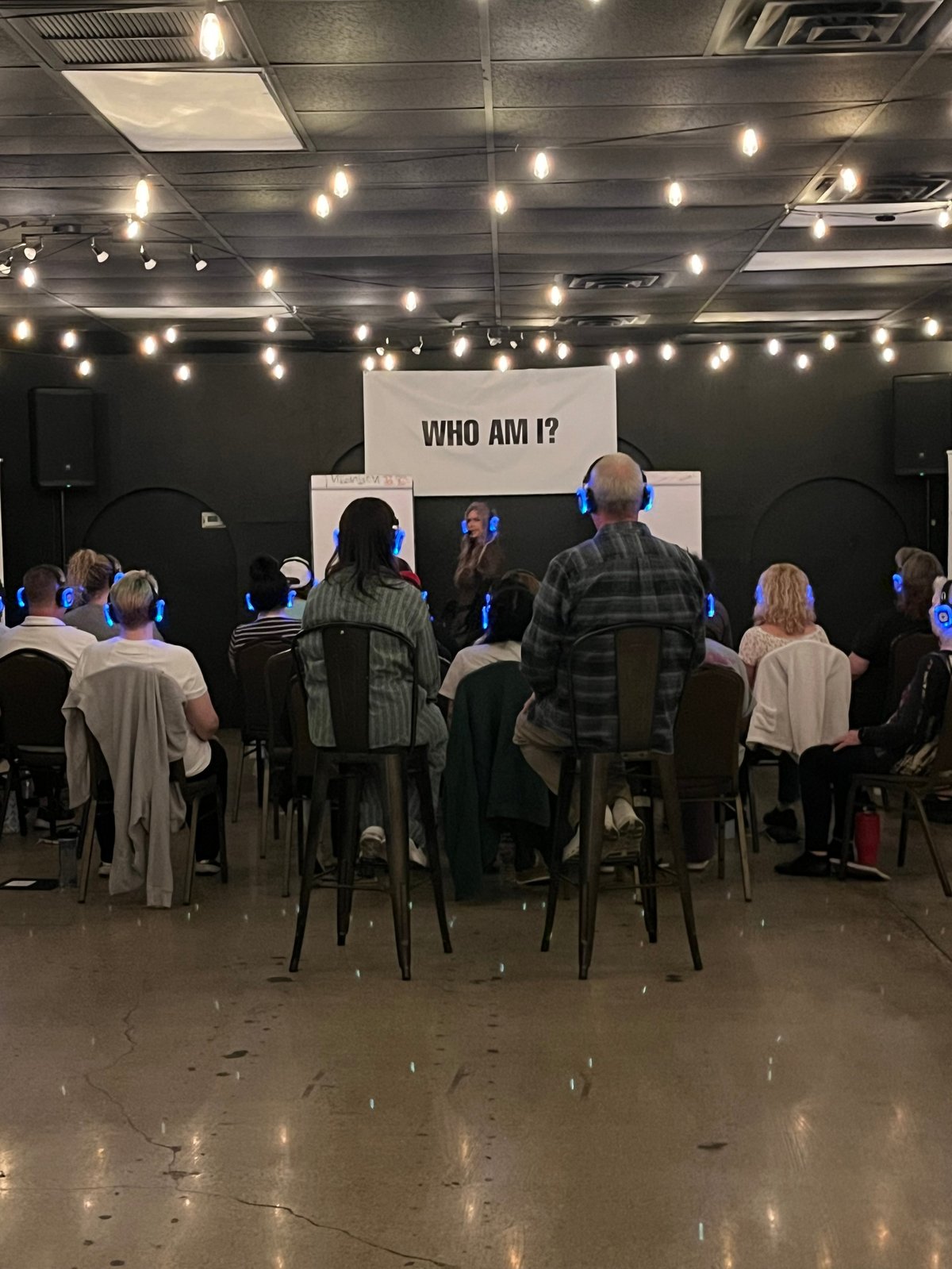 Group of people seated facing a stage with WHO AM I? sign at an indoor event with string lights overhead