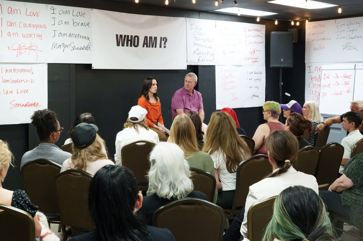 Two speakers lead a workshop titled WHO AM I? with audience members seated facing a stage with whiteboards displaying notes.