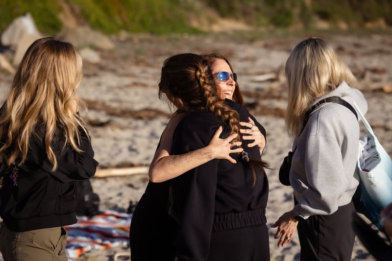 Three women with long hair hugging on a beach, wearing casual clothing and sunglasses
