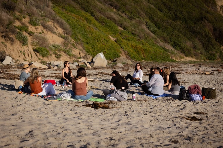 Group of people gathered on a sandy beach near a rocky cliff, sitting and socializing together