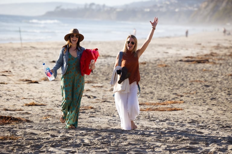 Two women walking on a beach, one wearing a cowboy hat and teal clothing, the other in a burgundy top with arm raised, waving