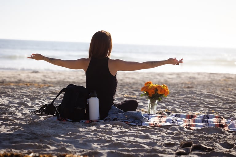 Woman sitting on beach with arms outstretched, flowers and picnic items nearby, ocean in background
