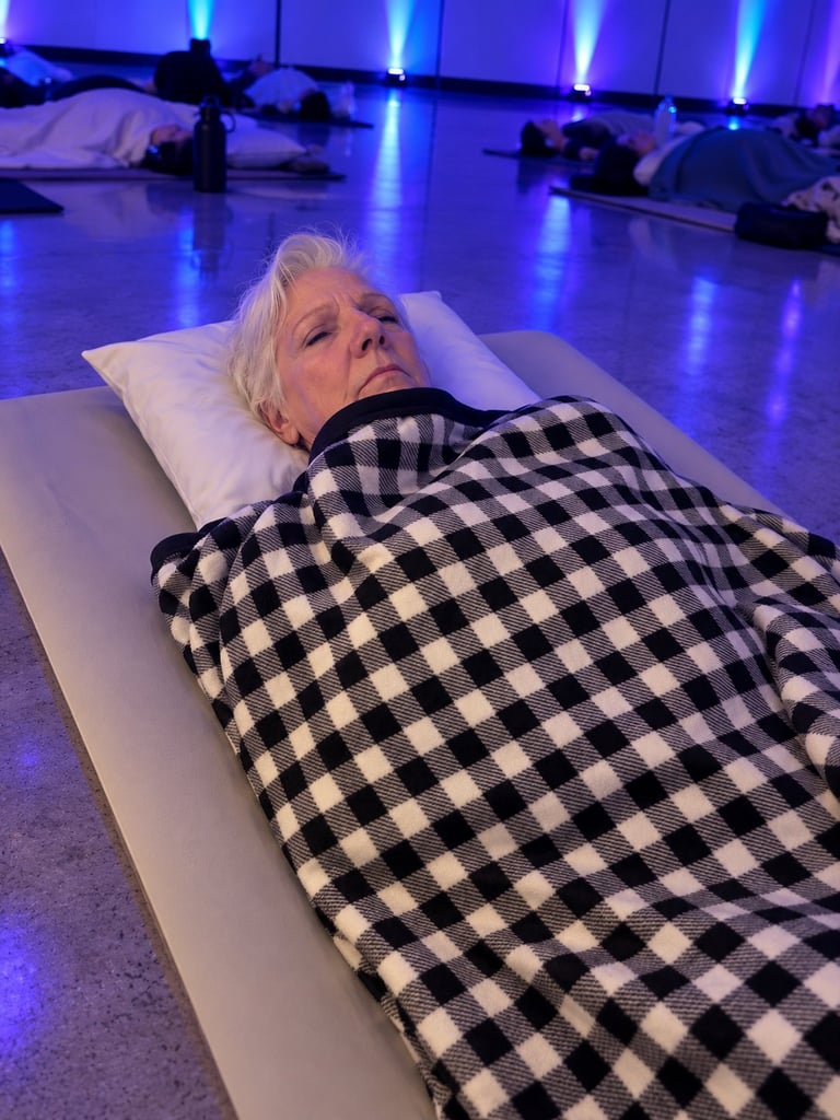 Woman with white hair lying on a white mat under a black and white checkered blanket in a purple-lit room with other people lying in background