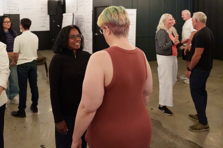 Group of people gathered indoors in a casual social setting, conversing in small clusters in what appears to be a community or event space.