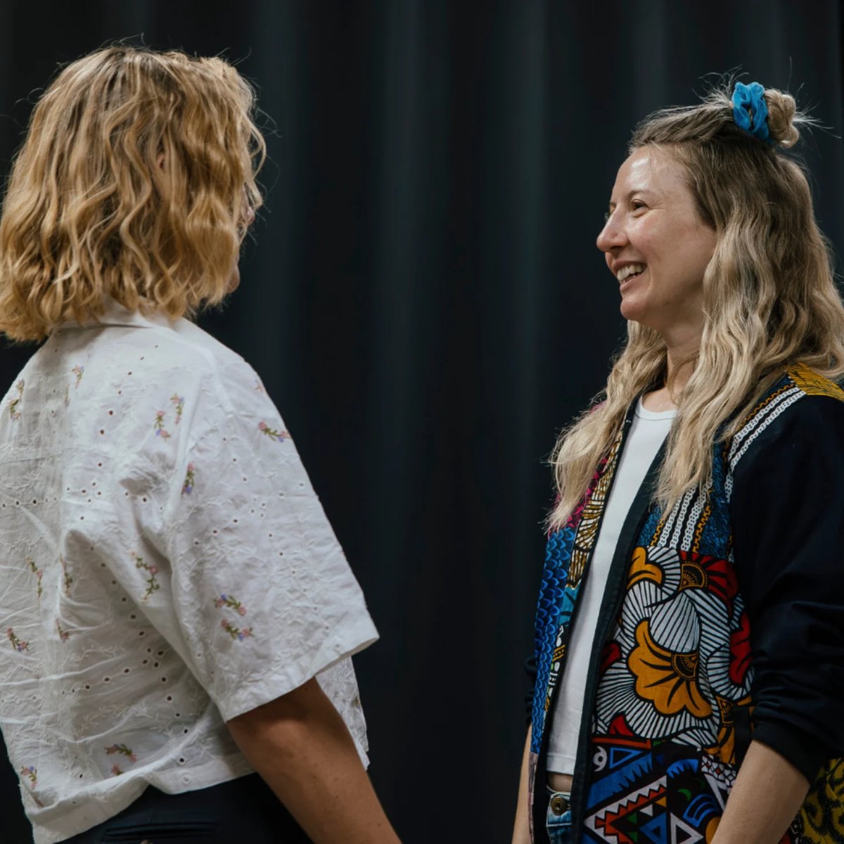 Two young women facing each other and smiling against a dark background, one wearing a white speckled shirt and the other wearing a colorful patterned jacket with a blue hair clip.