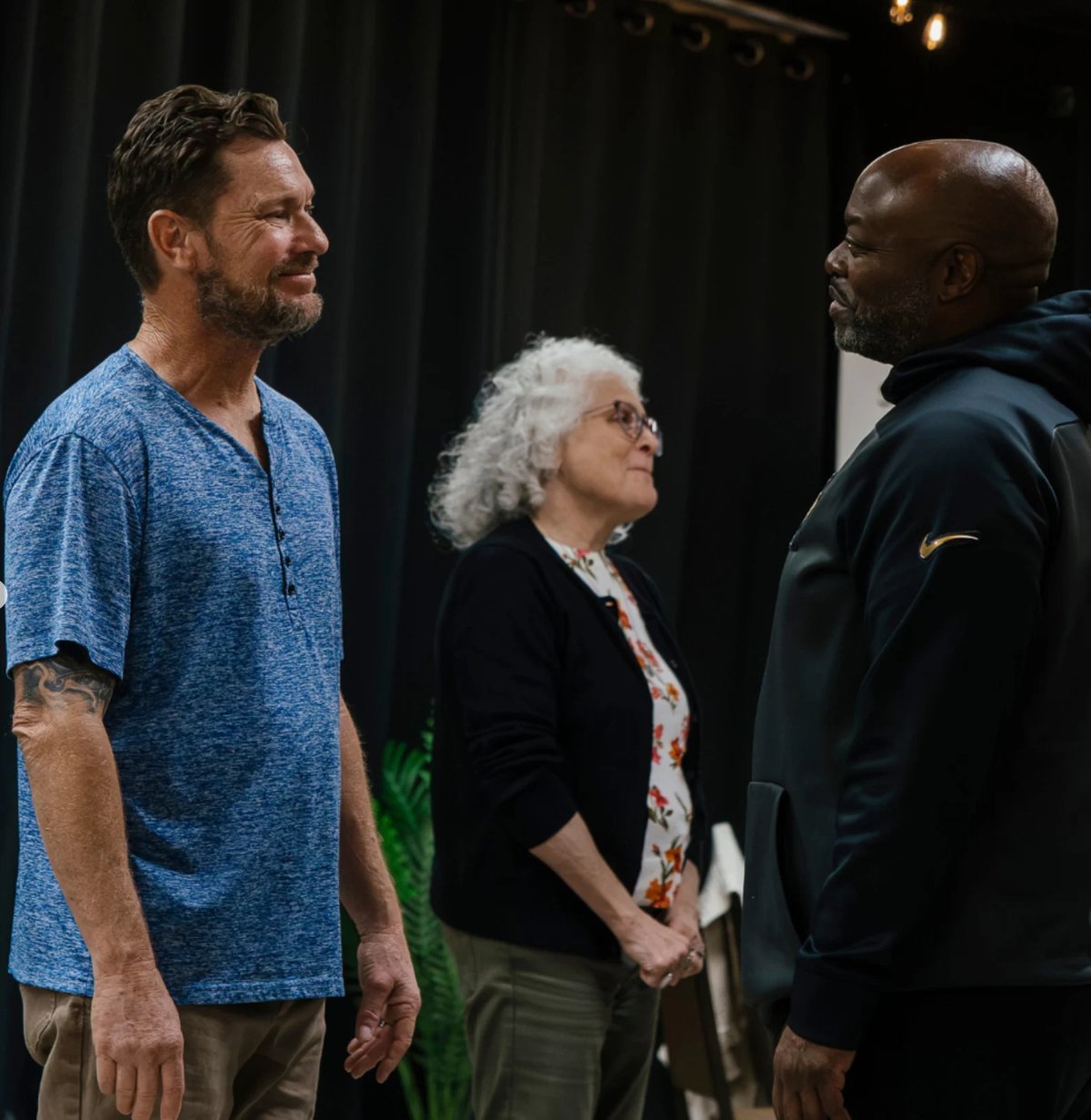 Three people having a conversation on stage: a man in a blue shirt on the left, an older woman with white hair in the middle, and a man in a black jacket on the right.