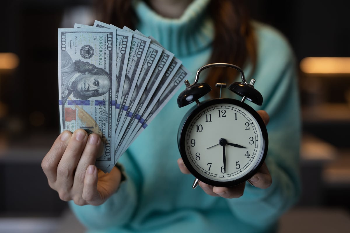 Person in teal shirt holding fanned dollar bills in one hand and a black alarm clock in the other, representing time and money concept