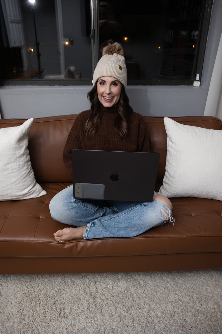 Woman sitting on brown leather couch wearing white beanie and brown sweater, working on laptop