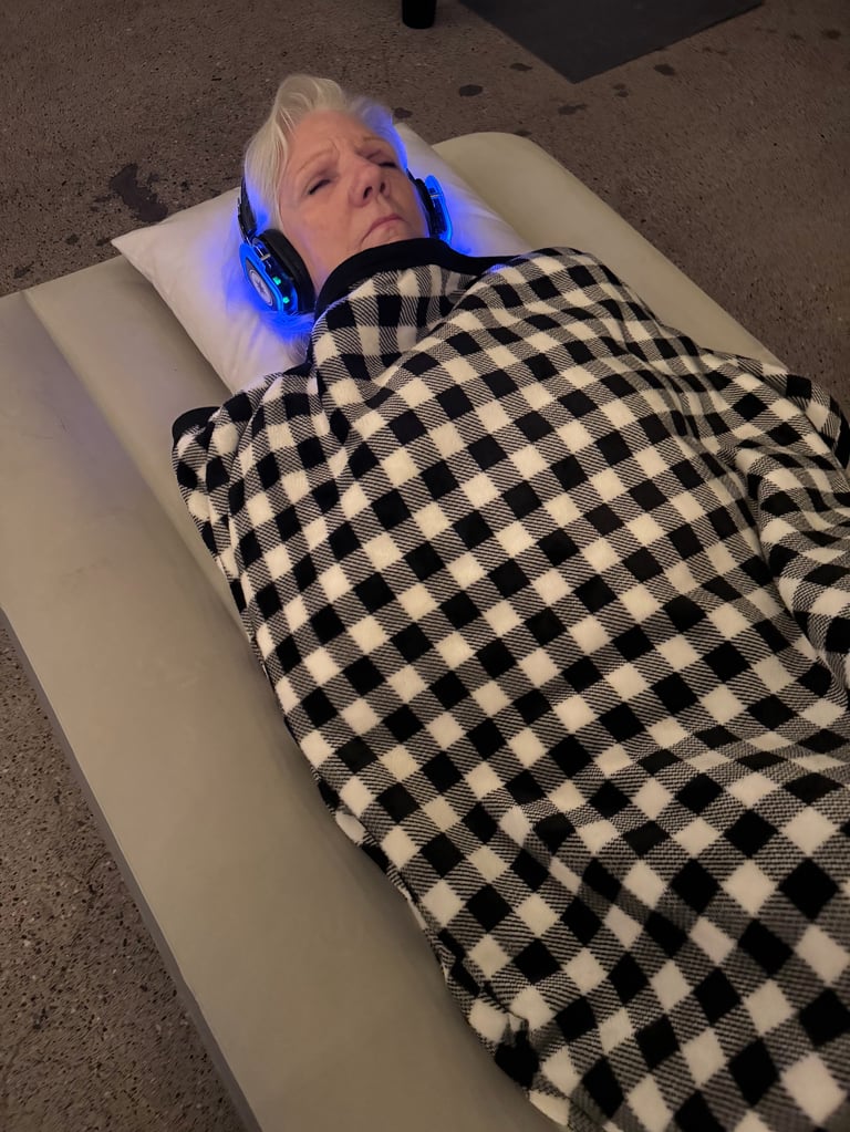 Person lying on white table receiving LED light therapy treatment with blue headgear and black and white checkered blanket