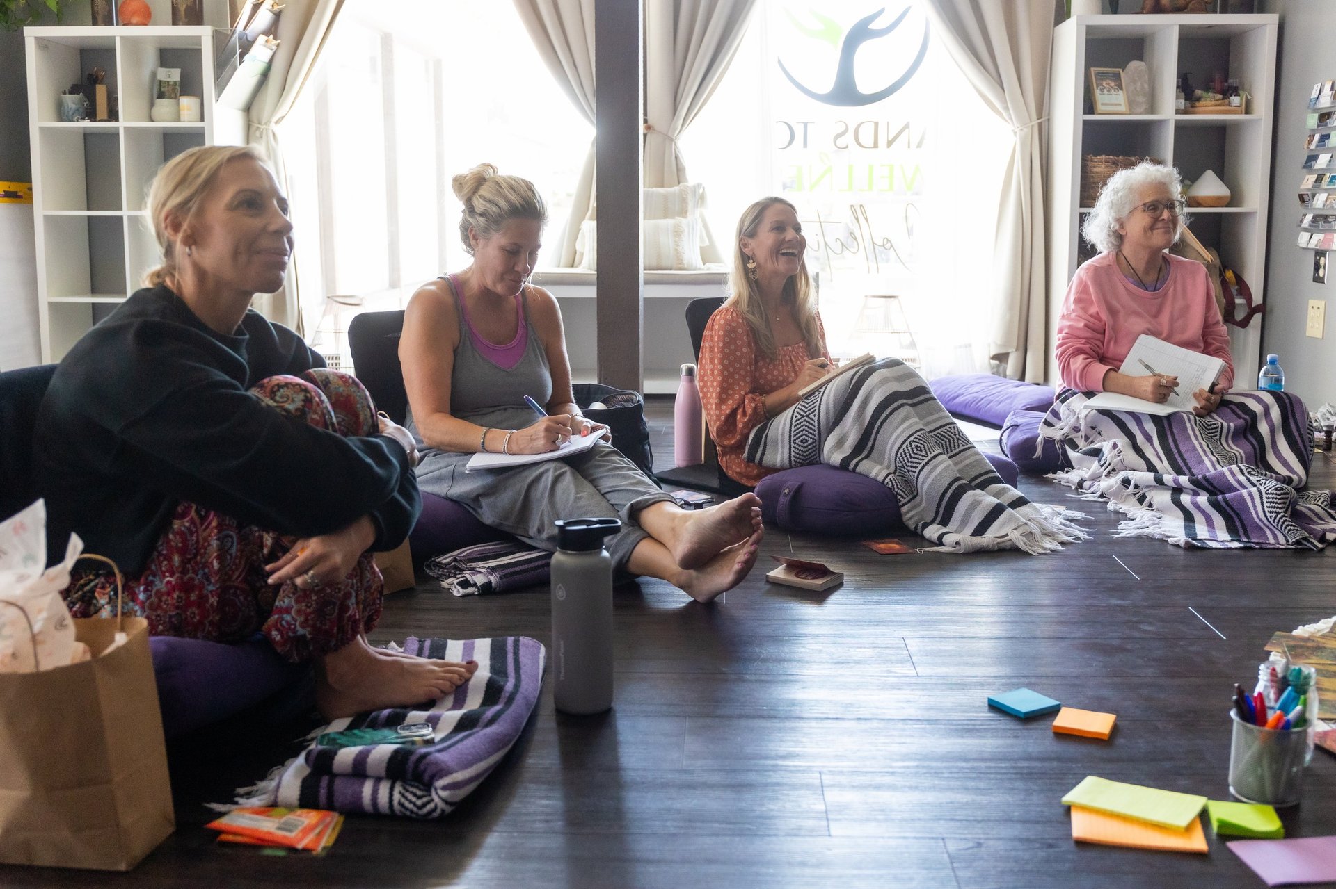Four women sitting on a studio floor organizing and folding clothing items during what appears to be a workshop or community event.