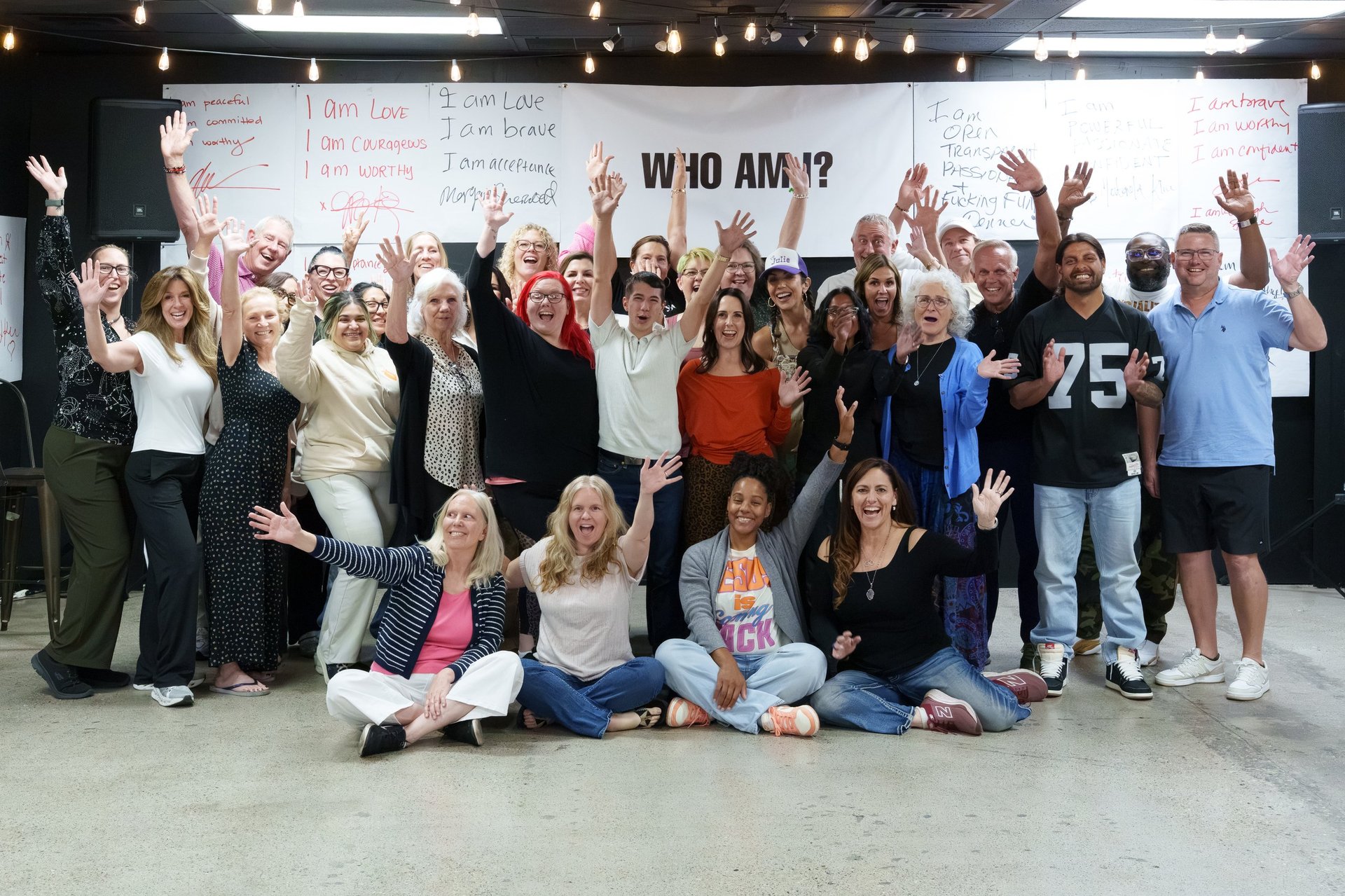 Large diverse group of people posing together with raised hands in front of a whiteboard that says WHO AM I? during a workshop or training session