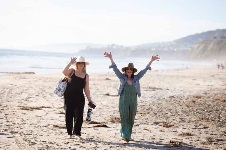 Two women walking on a sandy beach with arms raised, enjoying the seaside with cliffs in the background