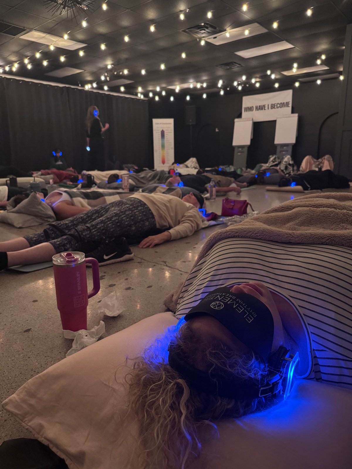 Group of people lying on the floor during a sound bath meditation session in a dimly lit room with string lights overhead