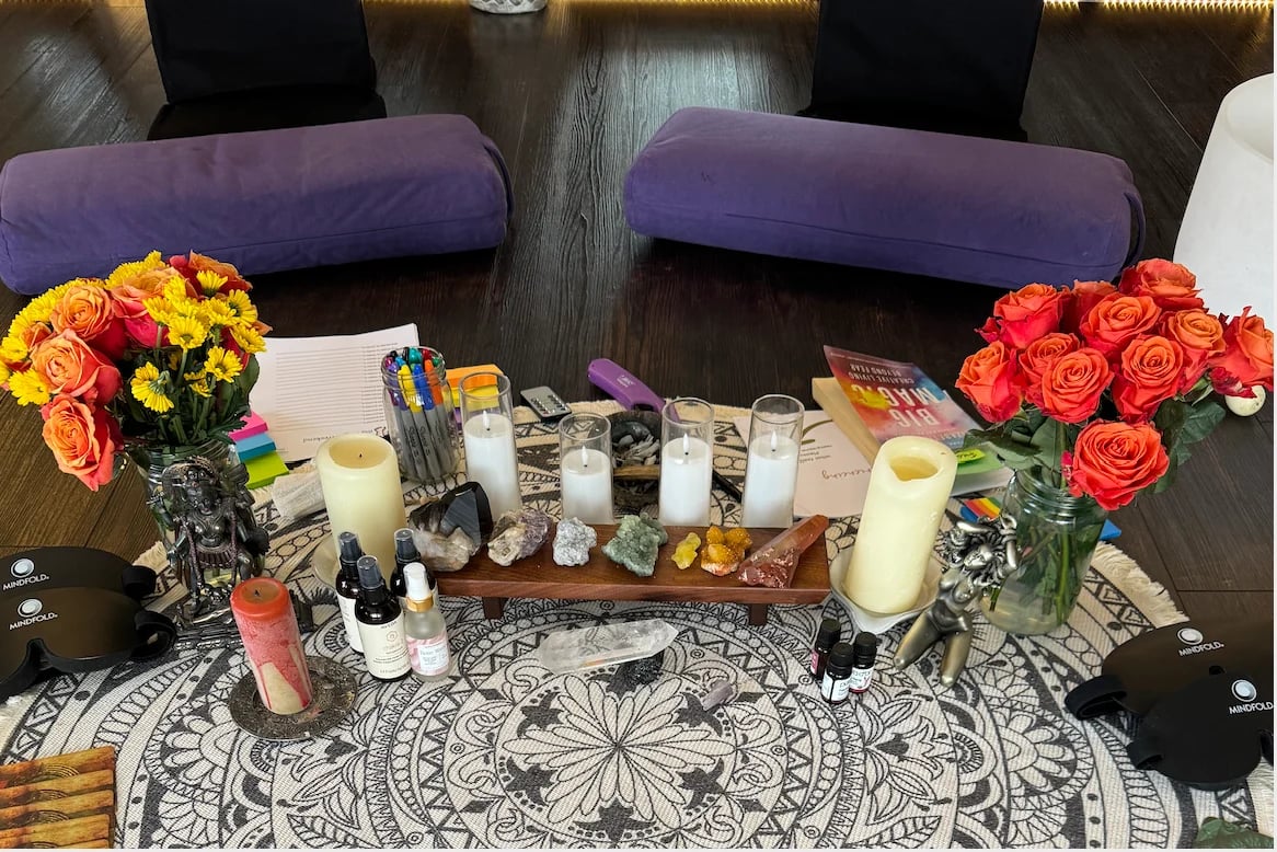 An altar display with colorful flower arrangements, lit candles, crystals, and decorative items on a patterned cloth with purple chairs behind