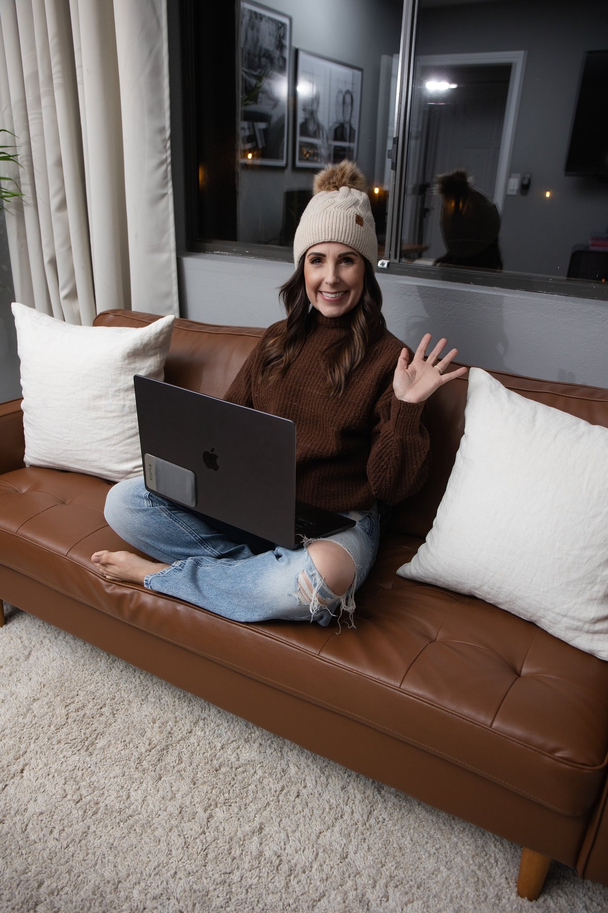 Woman sitting on a brown leather couch wearing a cream beanie and brown sweater, using a laptop and waving at the camera