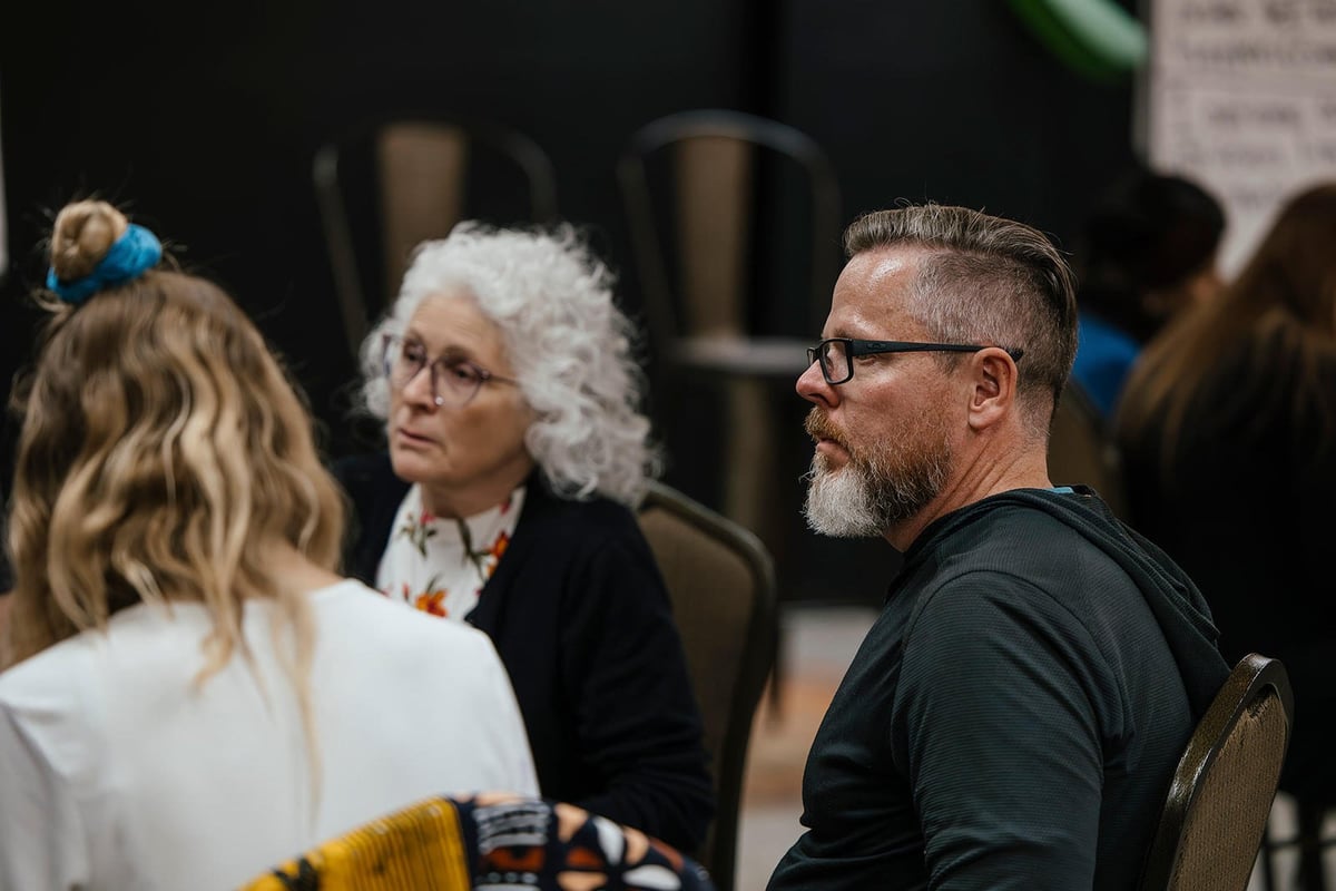 Group of people in conversation at an indoor event, including a man in glasses and black shirt speaking with others nearby